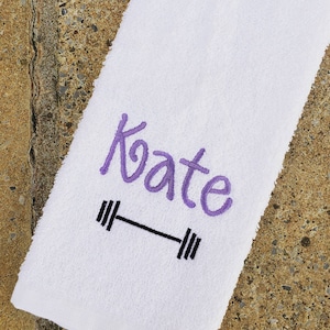 Puede incluir: Toalla blanca con el nombre "Kate" bordado en morado y un diseño de barra de pesas negra.