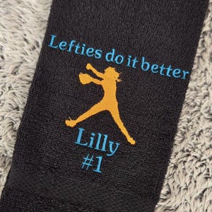 May include: Black towel with the text "Lefties do it better" in blue and a yellow silhouette of a baseball player. The text "Lilly #1" is also embroidered in blue.