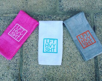 May include: Three towels with the text "LFT HVY SHT" embroidered in different colors. The towels are pink, white, and gray. The text is in a white square on the pink towel, a turquoise square on the white towel, and an orange square on the gray towel.