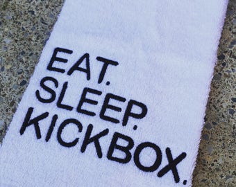 以下が含まれることがあります： 黒い刺繍で「EAT. SLEEP. KICKBOX.」と書かれた白いタオル