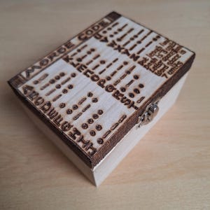 May include: A wooden box with a hinged lid and a metal clasp. The top of the box is decorated with a burned-in design of the Morse code alphabet and the text "MORSE CODE". The text below the code reads "Dot length is one unit, Dash is 3 units, Letter space is one unit, Word space is 7 units."
