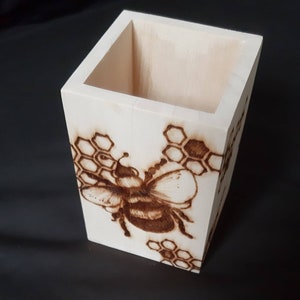 May include: A wooden pencil holder featuring a bee and honeycomb design. The holder is made from light wood and has a natural finish.