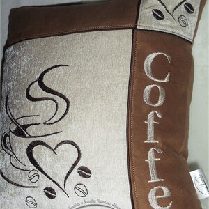 May include: A brown and beige throw pillow with the word "Coffee" embroidered in white on the right side. The left side features a stylized image of a coffee cup with steam and a heart, along with coffee beans. The pillow has a brown suede-like fabric and a beige fabric with a subtle texture.