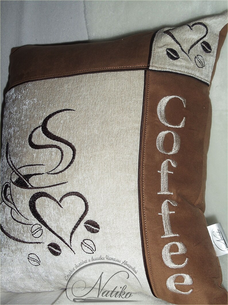 May include: A brown and beige throw pillow with the word "Coffee" embroidered in white on the right side. The left side features a stylized image of a coffee cup with steam and a heart, along with coffee beans. The pillow has a brown suede-like fabric and a beige fabric with a subtle texture.