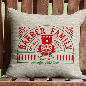 May include: A square, beige pillow with a holiday-themed design. The design features the words "BARBER FAMILY" in a semi-circle, with "CRUSHING HOLIDAYS" below. A red nutcracker is in the center, with "SINCE 1987" and "Brooklyn, New York" below.