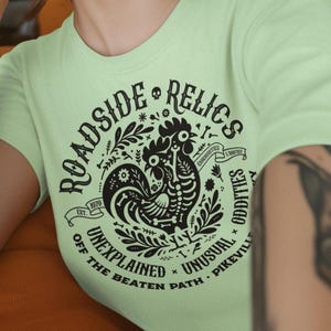 May include: A pale green t-shirt featuring a black graphic with the words "Roadside Relics" and a rooster design. The shirt also includes the phrases "Unexplained, Unusual, Off the Beaten Path, Pikeville" in a circular arrangement.