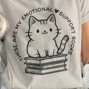 May include: A light beige t-shirt with a black and white illustration of a cat on books. The text reads "These are my emotional support books". The design is in a circular frame.