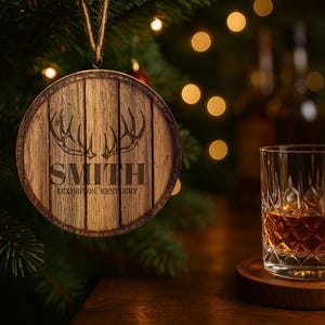 May include: A wooden Christmas ornament with the name "SMITH" and "Lexington, Kentucky" printed on it, hanging near a glass of amber liquid. The glass sits on a wooden coaster. The background features a Christmas tree and blurred bottles.