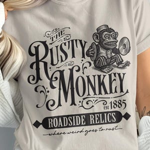 May include: Beige t-shirt featuring a vintage-style graphic. The design includes the text "The Rusty Monkey" with a monkey illustration holding a record, and the words "Roadside Relics" and "est 1885".