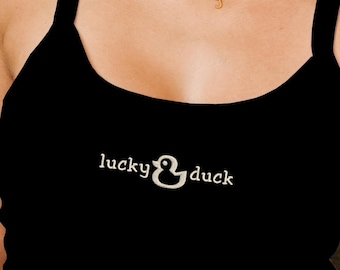 Puede incluir: Camiseta sin mangas negra con el texto "lucky & duck" en escritura blanca. La camiseta tiene un escote redondo y tirantes finos. La imagen muestra un primer plano de la camiseta.