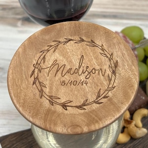 May include: A wooden wine glass topper with a wreath design and the name "Madison" and the date "5/10/14" engraved on it. The topper is sitting on top of a wine glass.