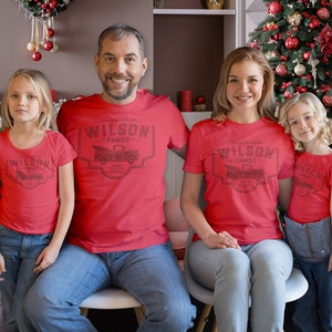 May include: A family of four wearing matching red t-shirts with a Christmas tree truck design and the text "Wilson Family Est. 2022". The family is standing in front of a decorated Christmas tree.