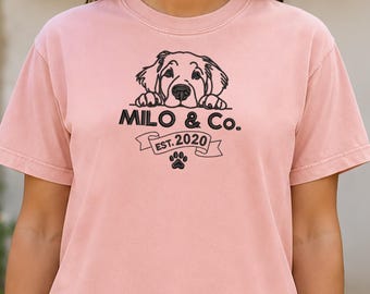 May include: A short-sleeved, crew-neck t-shirt in a soft peach color. The front features a black graphic illustration of a dog peeking over a banner with the text "MILO & Co." and "EST. 2020" above a small paw print icon centered on the chest.