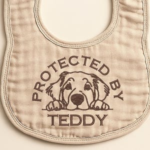 May include: A beige baby bib with a rounded shape and snap closures. The bib features a brown graphic of a dog and the text "PROTECTED BY TEDDY" in a circular design. The bib is made of a soft, textured fabric.