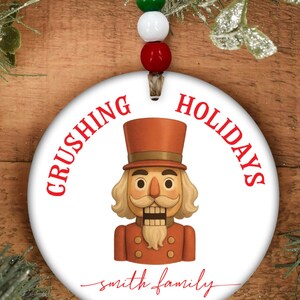 May include: A white, round Christmas ornament with the words "Crushing Holidays" in red. The ornament features a cartoon nutcracker in a brown uniform and top hat. The text "Smith Family Since 2019" is also included.