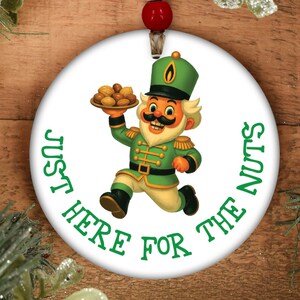 May include: White ceramic Christmas ornament with a cartoon nutcracker design. The nutcracker is wearing a green uniform and holding a plate of nuts. The text "JUST HERE FOR THE NUTS" is printed around the edge.