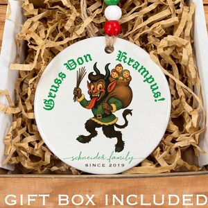 May include: A white ceramic Christmas ornament with a cartoon Krampus design, packaged in a gift box. The ornament features a green and black Krampus figure with the text "Gruss vom Krampus!" and "Schneider Family Since 2019". The ornament is strung with a beaded cord.