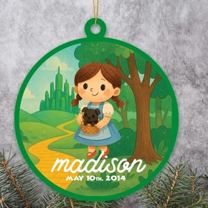 May include: A round, green Christmas ornament featuring a cartoon illustration of a girl in a blue dress holding a dog in a basket, with the text "madison MAY 10th, 2014". The background shows a yellow path, trees, and a city.