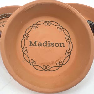 May include: Round terracotta-coloured trinket dish featuring the name "Madison" in black script within a decorative border. The dish has a slightly raised edge. Other similar dishes are visible.