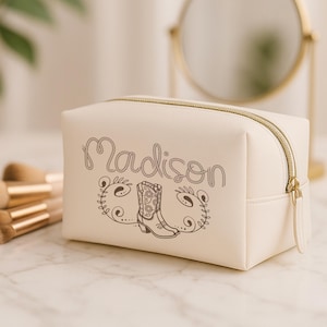 May include: Cream-colored cosmetic bag with a gold zipper and the name "Madison" in script. A decorative boot and floral design are printed on the front. Makeup brushes and a gold-framed mirror are in the background.