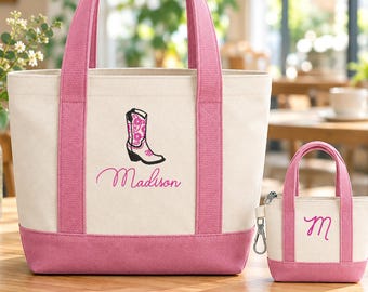 May include: A cream-colored canvas tote bag with pink handles and a pink base, featuring an embroidered pink cowboy boot and the name Madison in pink script. Beside it is a matching miniature canvas pouch with a pink letter M.