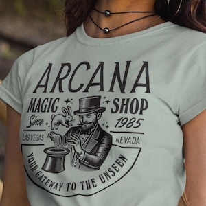 Puede incluir: Una mujer lleva una camiseta azul claro con un gráfico en blanco y negro de un mago con un sombrero de copa, un conejo y el texto "ARCANA MAGIC SHOP Since 1985 LAS VEGAS NEVADA YOUR GATEWAY TO THE UNSEEN". Lleva un sombrero de paja de ala ancha y pantalones cortos de mezclilla.