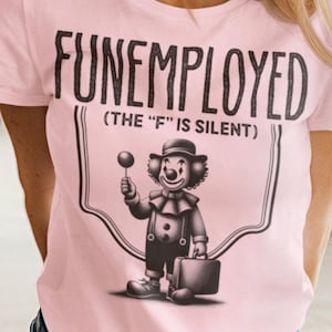 May include: A pink t-shirt with a black and white graphic of a clown holding a lollipop and a suitcase. The text on the shirt reads "FUNEMPLOYED (THE "F" IS SILENT)"