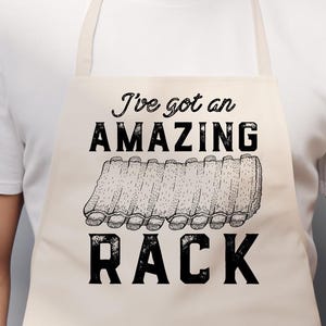 May include: Beige apron with the text "I've got an AMAZING RACK" in black. The design includes a graphic of a rack of ribs. The apron is worn by a person wearing a white t-shirt.