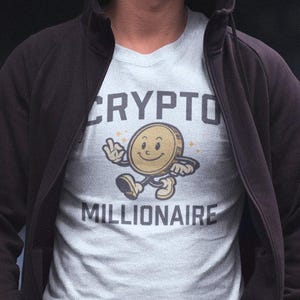 Op de afbeelding: Een persoon draagt een zwarte hoodie over een lichtblauw t-shirt met de tekst "CRYPTO MILLIONAIRE" en een cartoonafbeelding van een lachend muntpersonage.