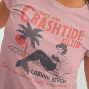 Make waves with the Crashtide Club tee, where vintage charm meets nautical grit. Featuring a sultry pinup mermaid reclining on a beach, this design oozes seaside rebellion. Faded ink and distressed lines give it that sun-washed, sea-worn look—perfect for beachcombers, rockabilly rebels, or anyone who sails by their own compass. Dive in. Danger and beauty never looked so good.