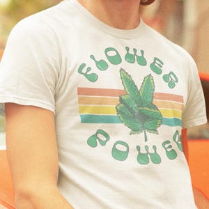 May include: A white t-shirt with a retro-style graphic that says "Flower Power" with a green marijuana leaf. The shirt is being worn by a person with curly hair and sunglasses.
