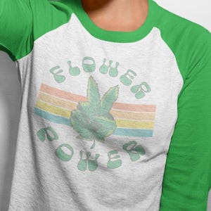 Peut inclure: Un t-shirt de style baseball blanc et gris avec des manches vertes. Le t-shirt présente les mots "Flower Power" en vert clair, avec un signe de paix et un motif arc-en-ciel.