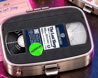 May include: A rectangular metal pill case featuring a black VHS tape design on the lid with the text The Lost Sleep and a green sticker labeled HORROR. The case is shown both closed and open, revealing two white interior compartments with pills.