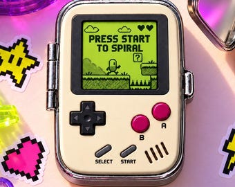 May include: A cream-colored pill case designed like a handheld video game console with a screen displaying the text PRESS START TO SPIRAL. The case is open, revealing four compartments containing small, round, and oval-shaped multi-colored pills.