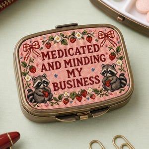Può includere: Un portapillole rettangolare in metallo con un coperchio ricamato rosa raffigurante due procioni, fragole e il testo "MEDICATED AND MINDING MY BUSINESS". La custodia è chiusa accanto a un portapillole aperto contenente compresse rotonde.