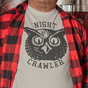 May include: A man wearing a red and black checked shirt over a white t-shirt with a black and white owl graphic and the text "NIGHT CRAWLER".