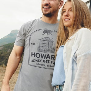May include: A couple standing in front of a white van, the man is wearing a grey t-shirt with the text "Howard Honey Bee Farm Rogers, Texas" printed on it, the woman is wearing a white shirt over a blue shirt and jeans.