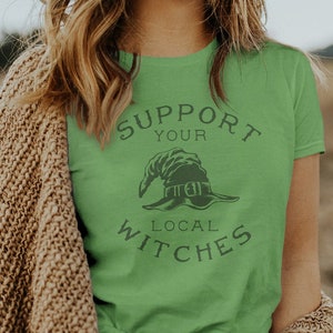 Pode incluir: Uma mulher vestindo uma camiseta verde com o texto "Support Your Local Witches" e um gráfico de um chapéu de bruxa. Ela está vestindo um cardigã marrom e um cinto preto com uma fivela de prata.