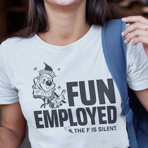 May include: White t-shirt with the words "FUN EMPLOYED" in bold black letters and a cartoon jester graphic. The text "THE F IS SILENT" is below the main text. The shirt is being worn by a person with a blue backpack.