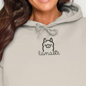 Op de afbeelding: Een lichtbeige hoodie met een voorzak en een capuchon met trekkoord. De hoodie heeft een kleine zwarte afbeelding van een lama met het woord "lamaste" in script eronder. De hoodie is gemaakt van een zacht materiaal.