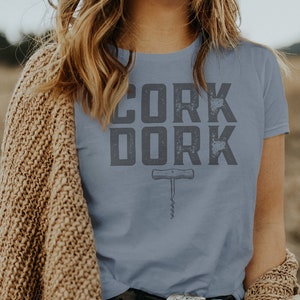May include: A woman wearing a light blue t-shirt with the text "CORK DORK" and a corkscrew graphic. She is wearing a brown cardigan and black jeans.