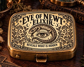 May include: A rectangular brass-toned metal tin featuring an engraved design of a lizard curled around an eye. The text reads EYE OF NEWT ELIXIR and REVEALS WHAT IS HIDDEN. The lid is decorated with intricate floral scrollwork and a latch closure.