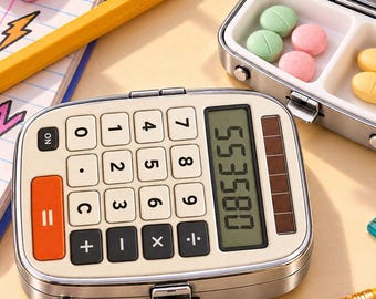 May include: A silver metal pill case designed to look like a calculator with a digital display showing 553580, featuring a hinged lid that opens to reveal two compartments containing small round green, pink, yellow, and white pills.