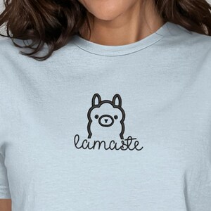 Op de afbeelding: Lichtblauw T-shirt met een zwarte lama-illustratie en het handgeschreven woord "lamaste". Het shirt is gemaakt van een zacht materiaal en is geschikt voor casual wear. Het ontwerp is eenvoudig en minimalistisch.
