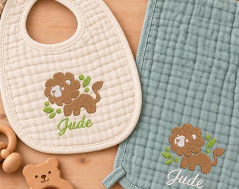 May include: A cream-colored quilted baby bib and a sage green quilted burp cloth, both featuring an embroidered brown lion and the name Jude. Also included are a pair of cream-colored knit baby booties and a light brown wooden bear-shaped rattle.