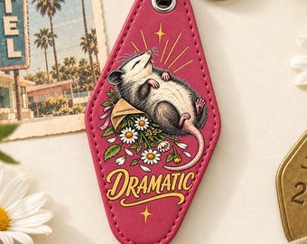 May include: A pink motel-style keychain featuring a graphic of a possum lying on its back amidst white daisies. The word DRAMATIC is printed in yellow cursive text at the bottom. The keychain includes a black metal connector and a silver ring.