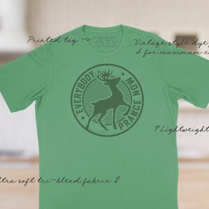 Op de afbeelding: Een groen T-shirt met een vintage stijl dye-print van een hert in een cirkel met de tekst "Everybody Mon Prance". Het shirt is gemaakt van een zachte tri-blend stof en heeft een lichtgewicht gevoel.