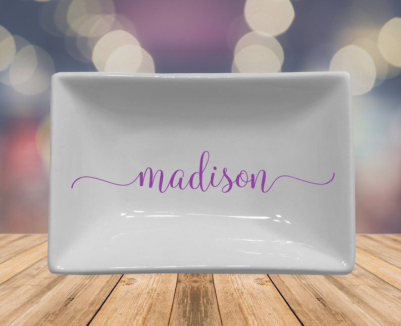 May include: A white rectangular ceramic dish with the name "madison" in purple script. The dish has a slightly raised edge and sits on a wooden surface. The background is blurred with bokeh lights.