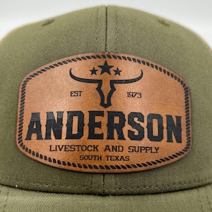 May include: Olive green and beige trucker cap with a leather patch. The patch displays a bull skull with stars, the name "ANDERSON", and the text "LIVESTOCK AND SUPPLY SOUTH TEXAS".