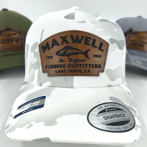 May include: A white camouflage baseball cap with a brown leather patch featuring the text "MAXWELL" and "FISHING OUTFITTERS LAKE TAHOE, CA". The cap has a curved brim and a snapback closure. The hat is a Yupoong product.